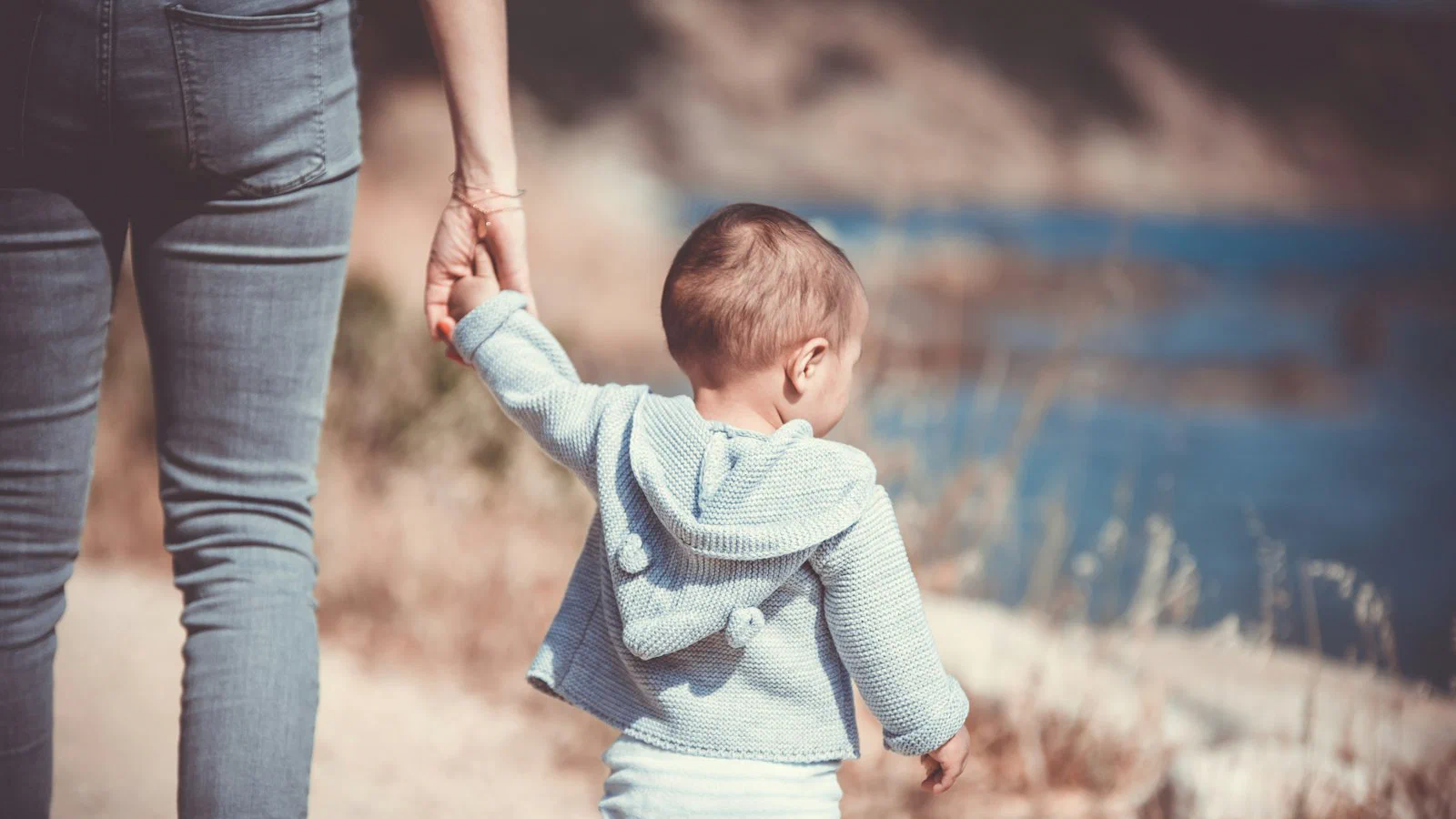Adulto e criança de mãos dadas, vistos de costas, caminhando por uma praia
